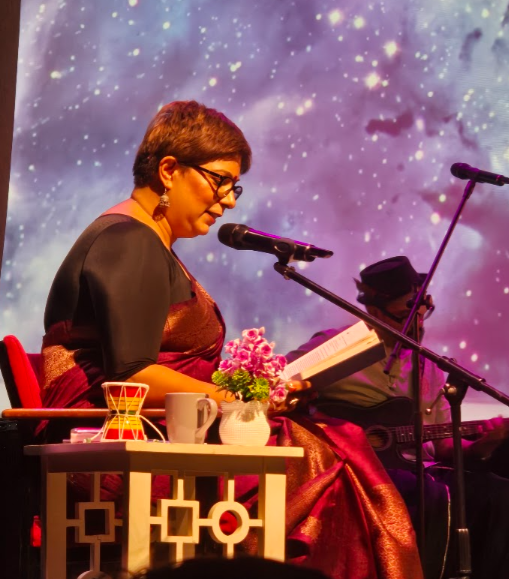 Lakshmi Kaul reading from Little Divine Ones on stage at The Bhavan in London, with a musician playing guitar behind her